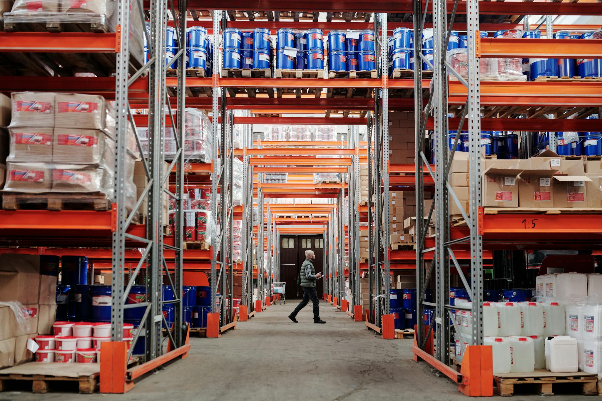 A man walking through a large industrial warehouse with stacked shelves filled with goods and products.